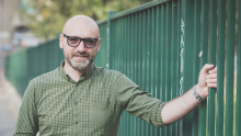 Man in khaki green shirt with sunglasses, leaning against a green park fence