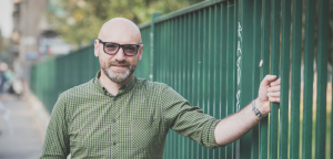 Man in khaki green shirt with sunglasses, leaning against a green park fence